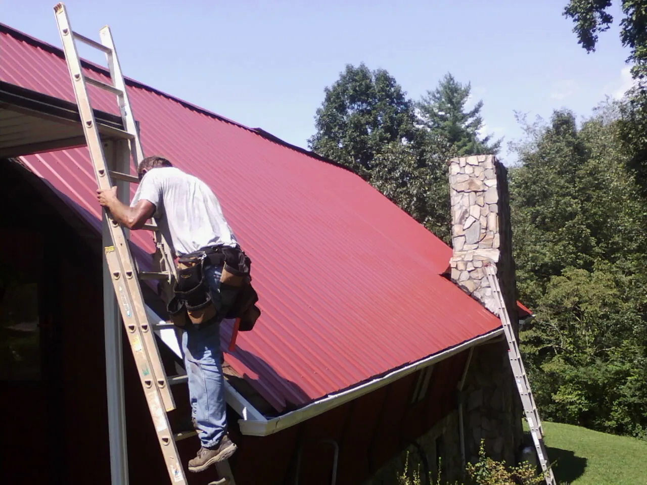 Roofer on a ladder beside a red metal roof and stone chimney — Soffit & Fascia Repair by Roofing Brothers in Sharpsburg